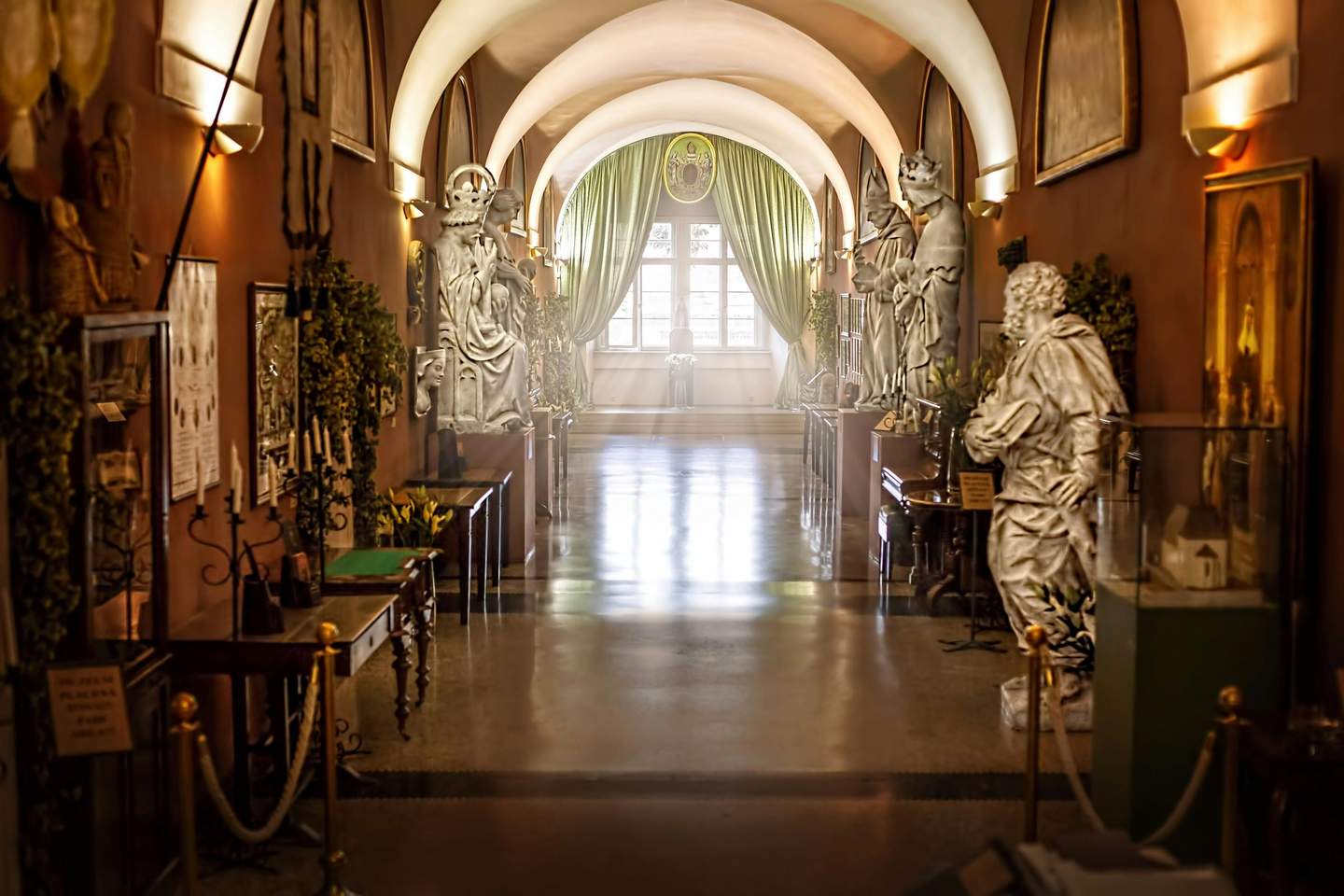 Charles Bridge Museum A museum hallway with statues, framed artworks, and display tables on both sides; illuminated by natural light from a window at the end.