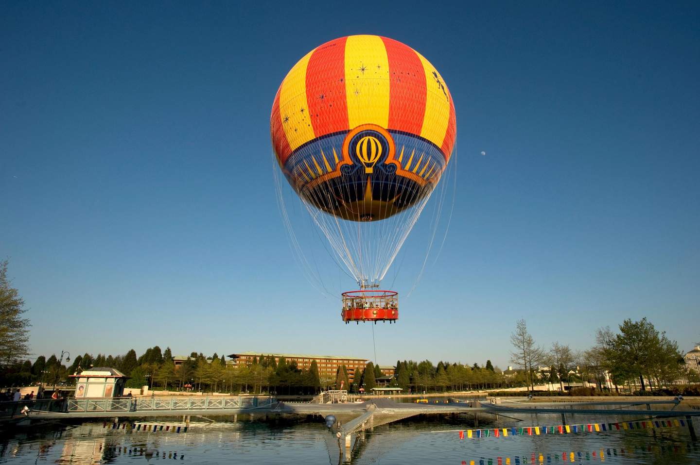 Paris Balloon Flights A colorful hot air balloon is tethered above a pond with a dock, surrounded by trees and buildings under a clear blue sky.