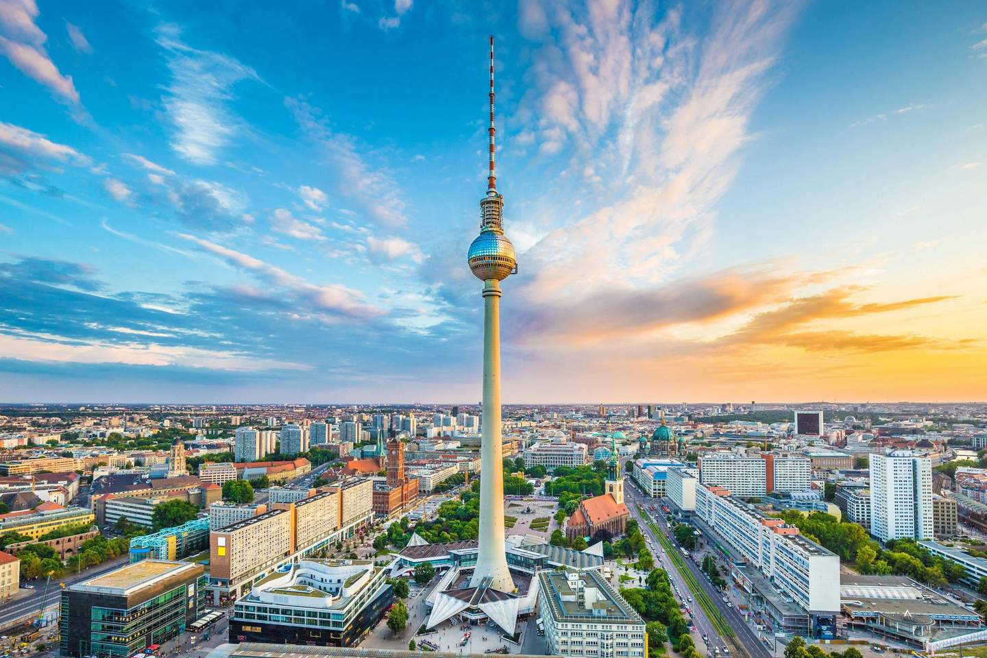 Aerial view of a cityscape featuring a tall TV tower at the center, surrounded by buildings, streets, and greenery under a blue sky.