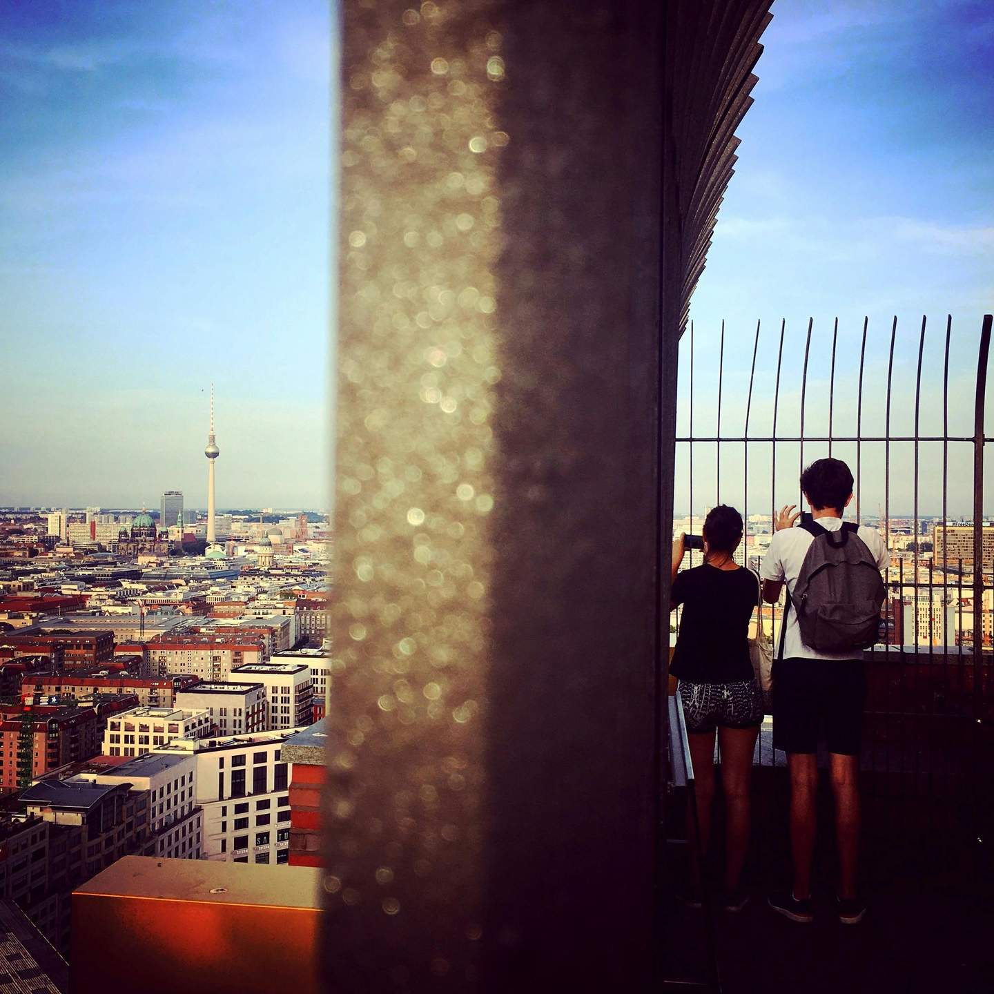 Two people viewing a cityscape from a high observation deck, separated by a vertical partition. The skyline includes a prominent TV tower.