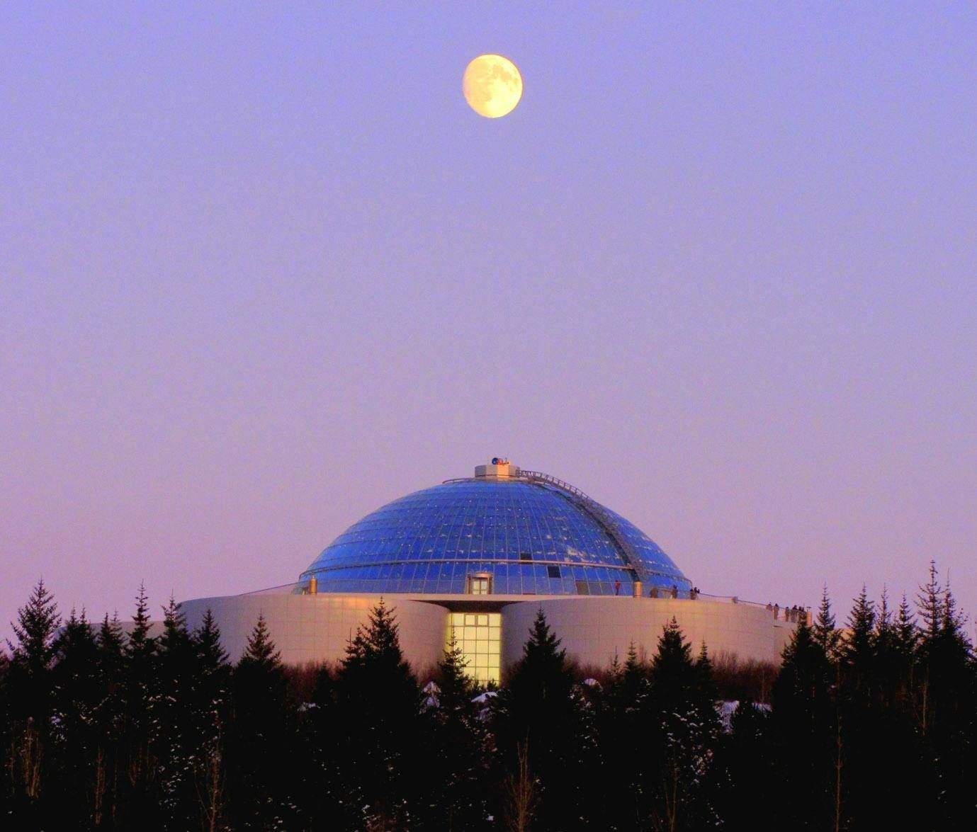 Perlan A glass-domed building surrounded by trees, with a full moon in a pink and blue sky above.