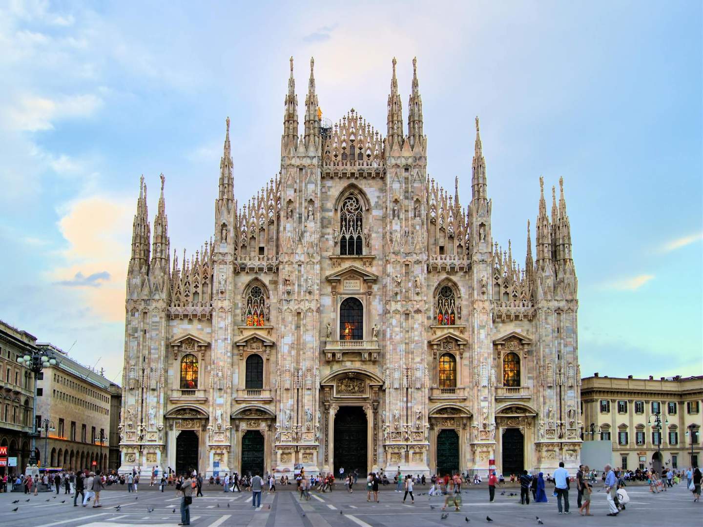Duomo di Milano People gather in front of an ornate Gothic cathedral with intricate spires and stained glass windows under a blue and cloudy sky.