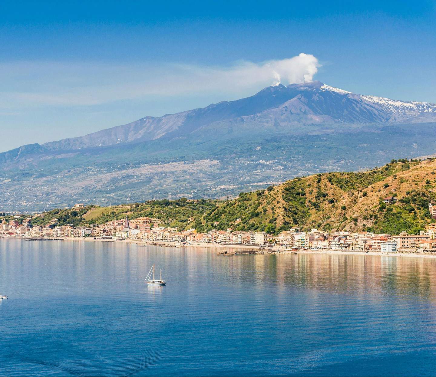 Mount Etna Coastal town with buildings and hills in foreground, a sailboat on calm water, and a smoking volcano in the background.