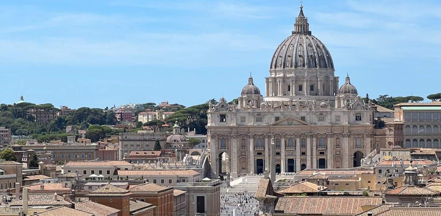 St. Peter's Basilica View of St. Peter's Basilica with its large dome, surrounded by rooftops and trees under a clear blue sky.