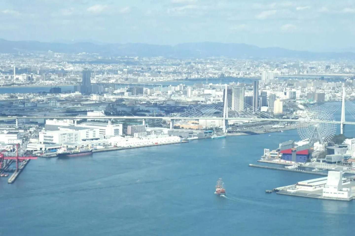 A cityscape with a large waterfront, a suspension bridge, a Ferris wheel, industrial buildings, and a ship in the water.