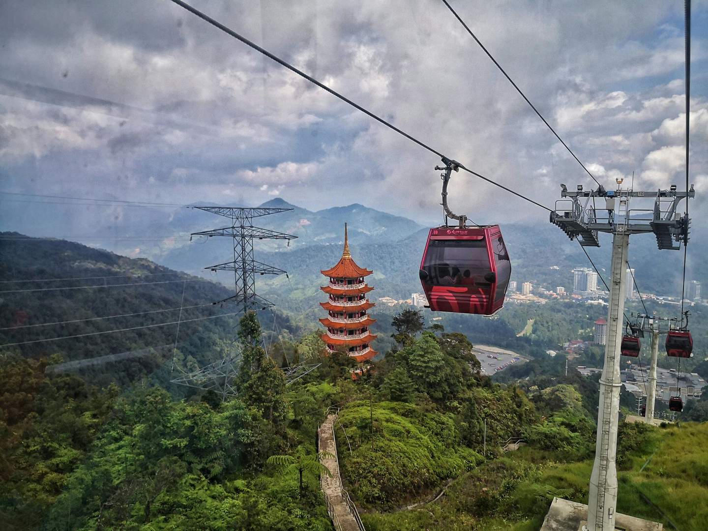 Genting Highlands Red cable cars travel over a lush mountain landscape with a multi-tiered pagoda and towering power lines, under a cloudy sky.