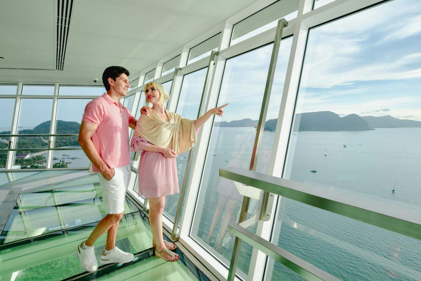A couple stands on a glass floor in a modern building, looking out at a scenic ocean view through large windows.