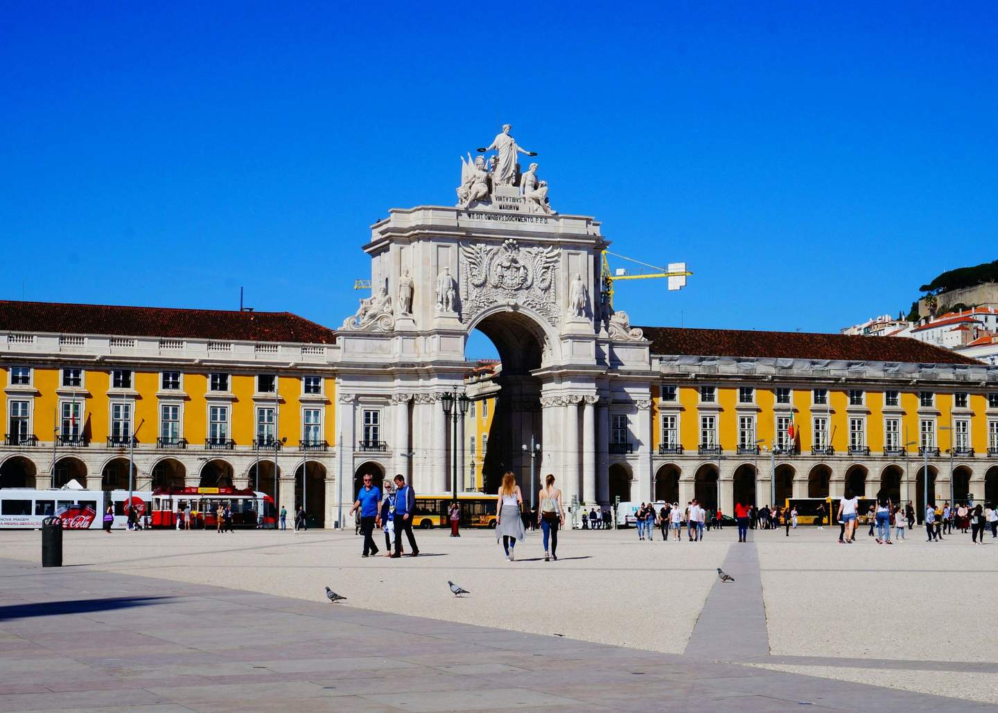 People walking in a large square with a yellow building and an ornate archway under a clear blue sky.