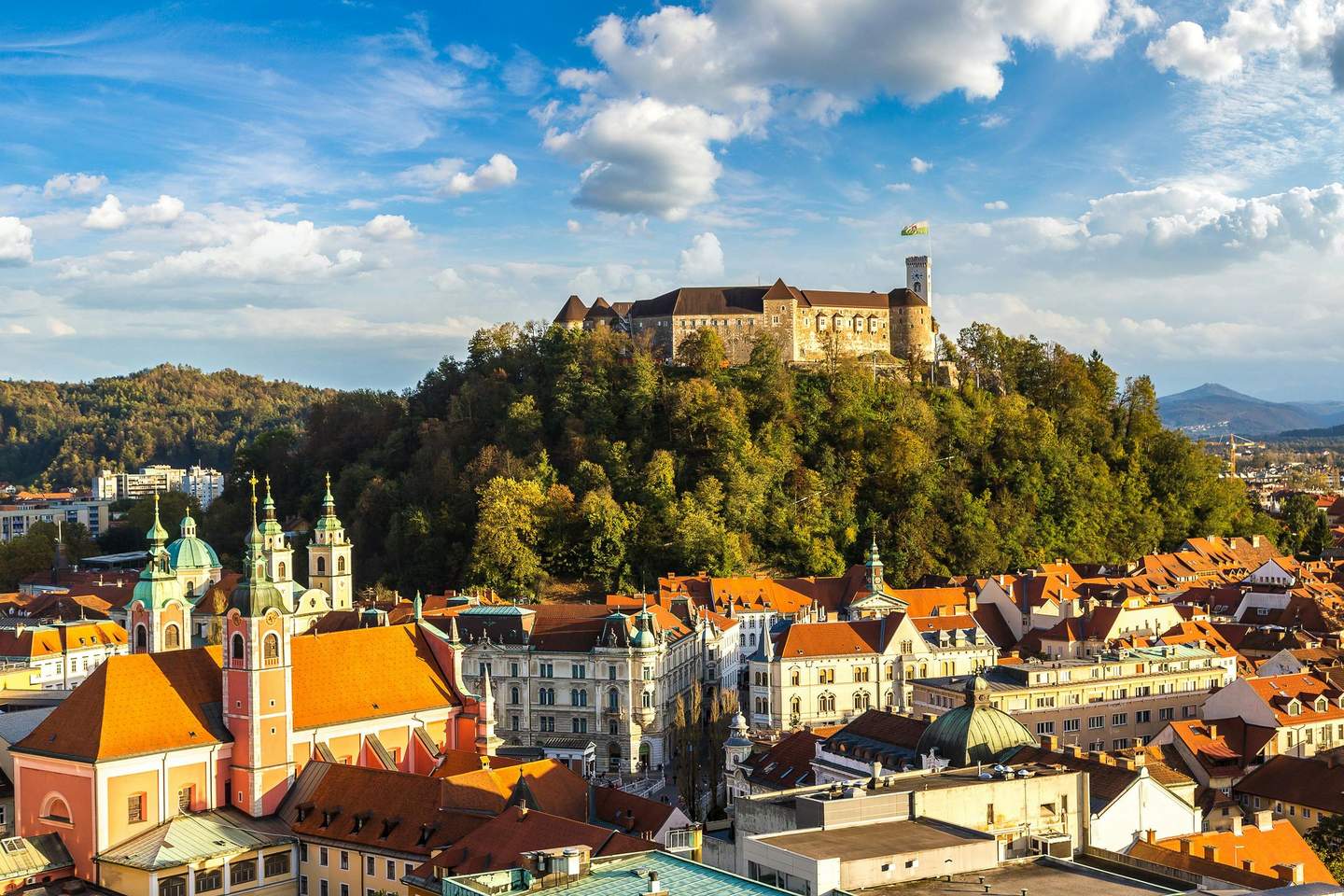 Ljubljana Castle A historic castle on a forested hill overlooks a cityscape with red-roofed buildings and church towers under a partly cloudy sky.