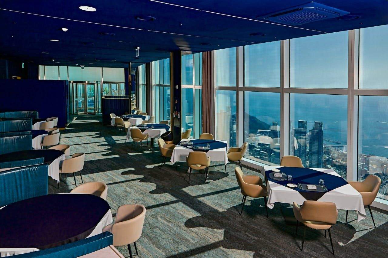 A modern dining area with large windows overlooking a cityscape and ocean, featuring tables with white and blue tablecloths.