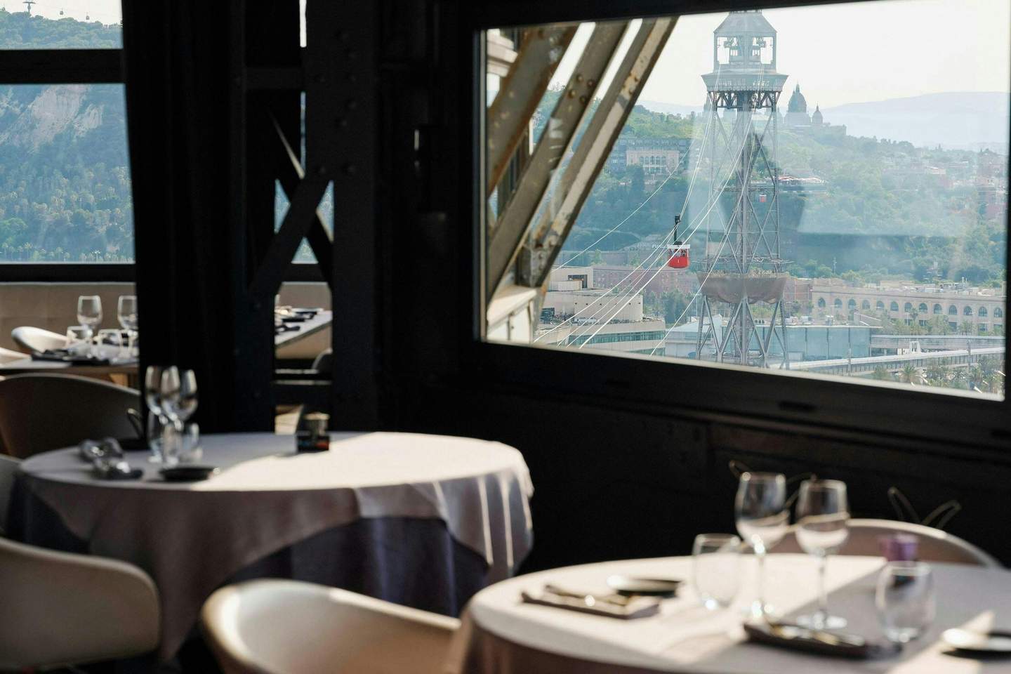Elegant dining tables with glasses and utensils next to a window showcasing a cityscape with a cable car and mountains in the background.