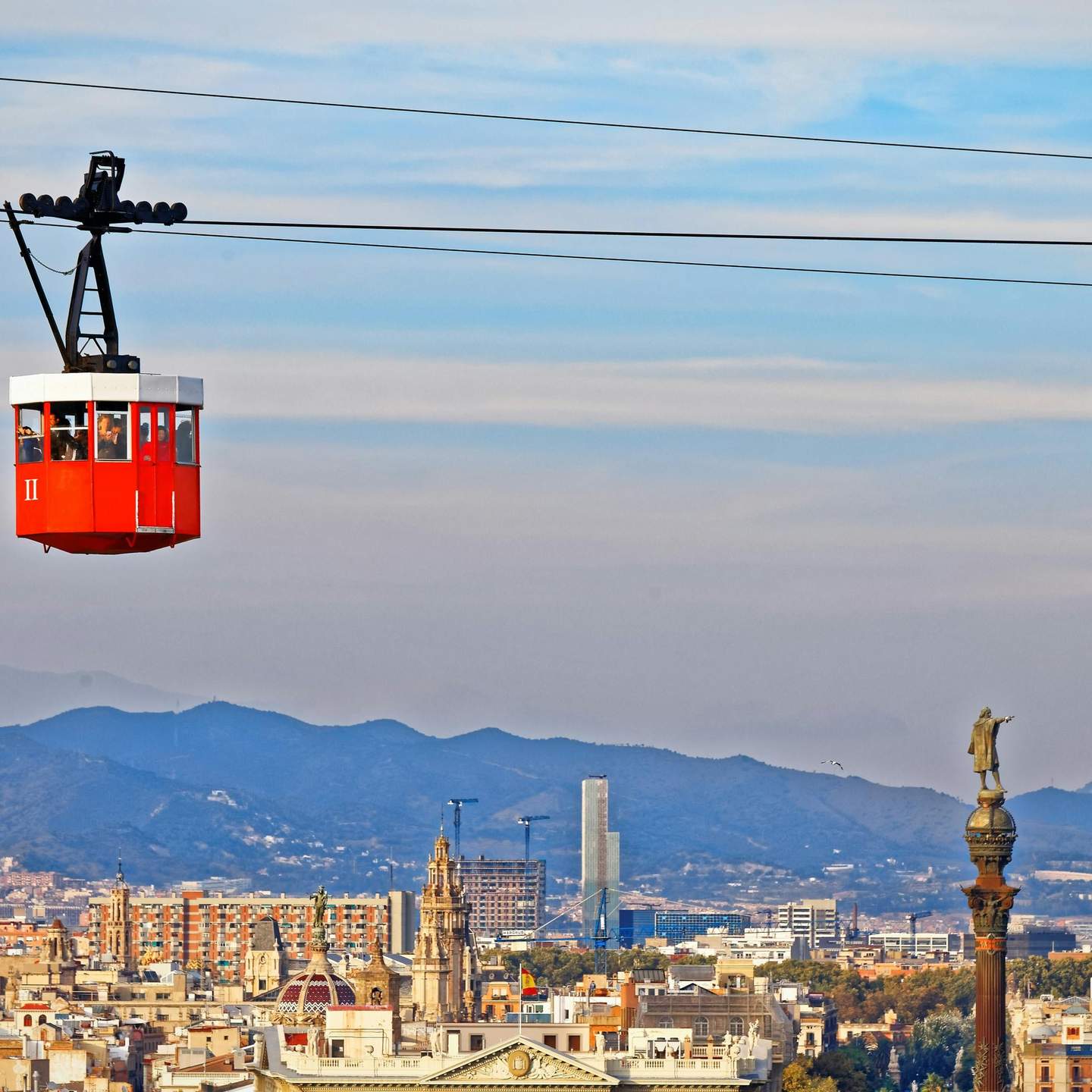 Barcelona Cable Car (Port Cable Car) A red cable car travels across the sky with a cityscape and mountains in the background under a cloudy sky.