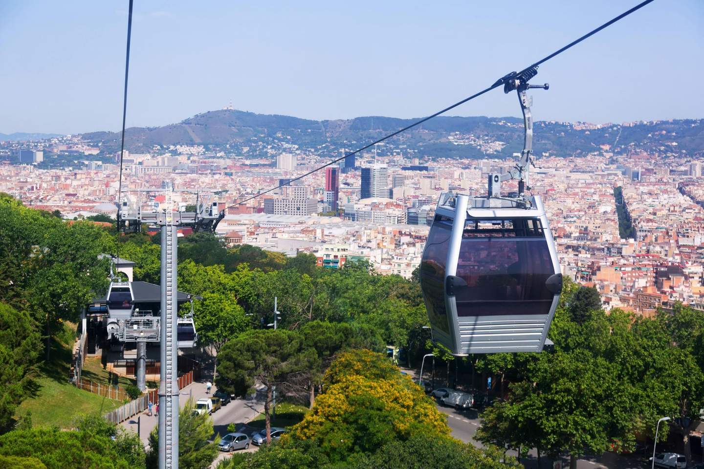 Montjuïc Cable Car Cable cars moving above lush greenery with a densely packed cityscape and mountains in the background.