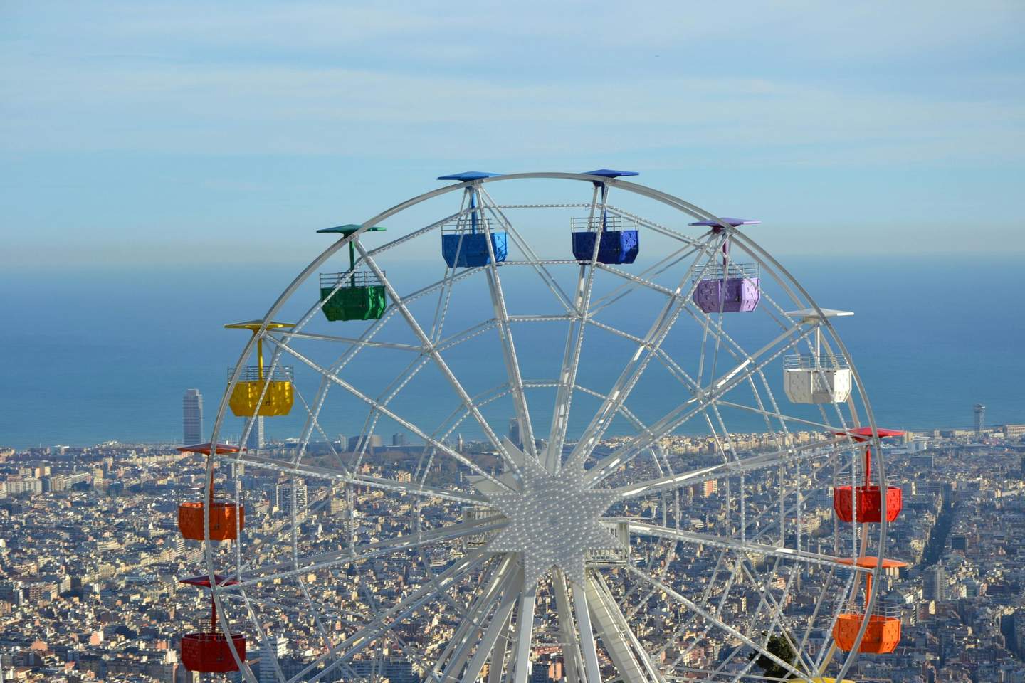 Ferris wheel with multicolored cabins overlooks a cityscape and ocean under a clear sky.