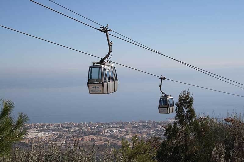 Teleférico Benalmádena Two cable cars travel over a hilly landscape with a coastal city and ocean in the background on a clear day.