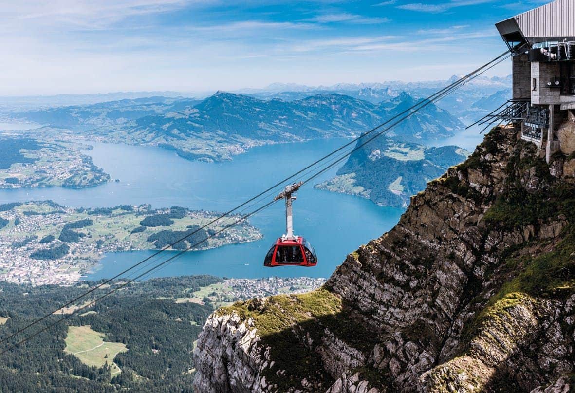 Dragon Ride (Pilatus Kulm) A red cable car travels over a steep mountain cliff with a scenic view of a lake and distant green hills below.
