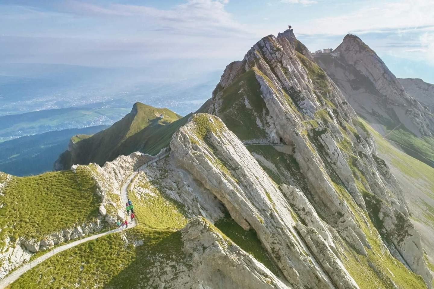 Hikers walking on a narrow mountain trail with steep, rocky peaks nearby and a vast landscape in the distance.