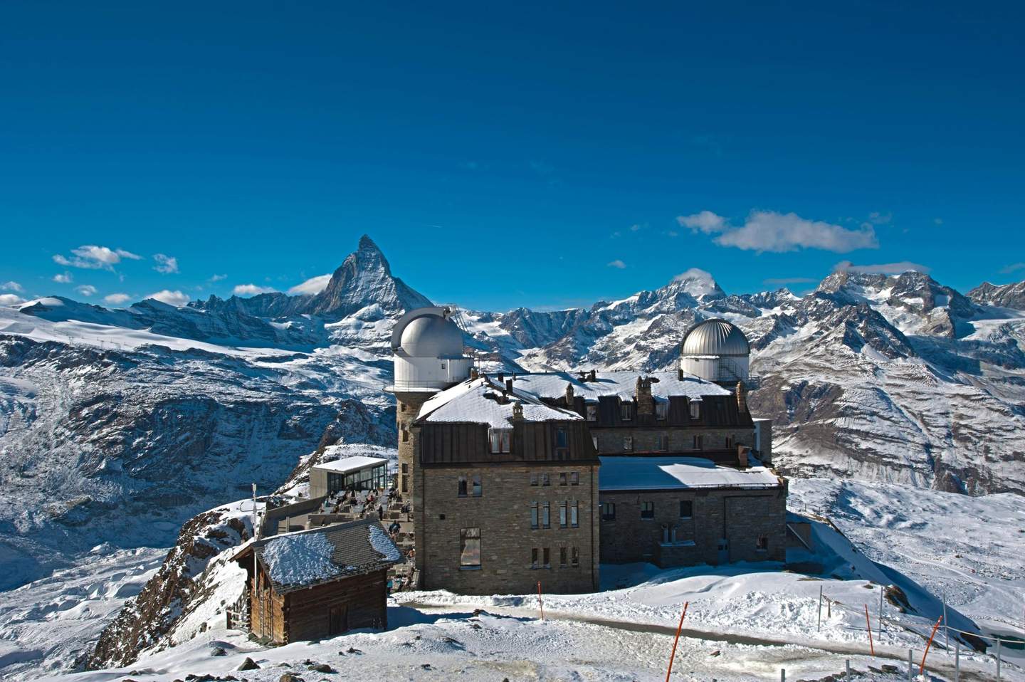Gornergrat A snow-covered observatory with two domes on a mountain, with the Matterhorn and surrounding peaks under a clear blue sky.