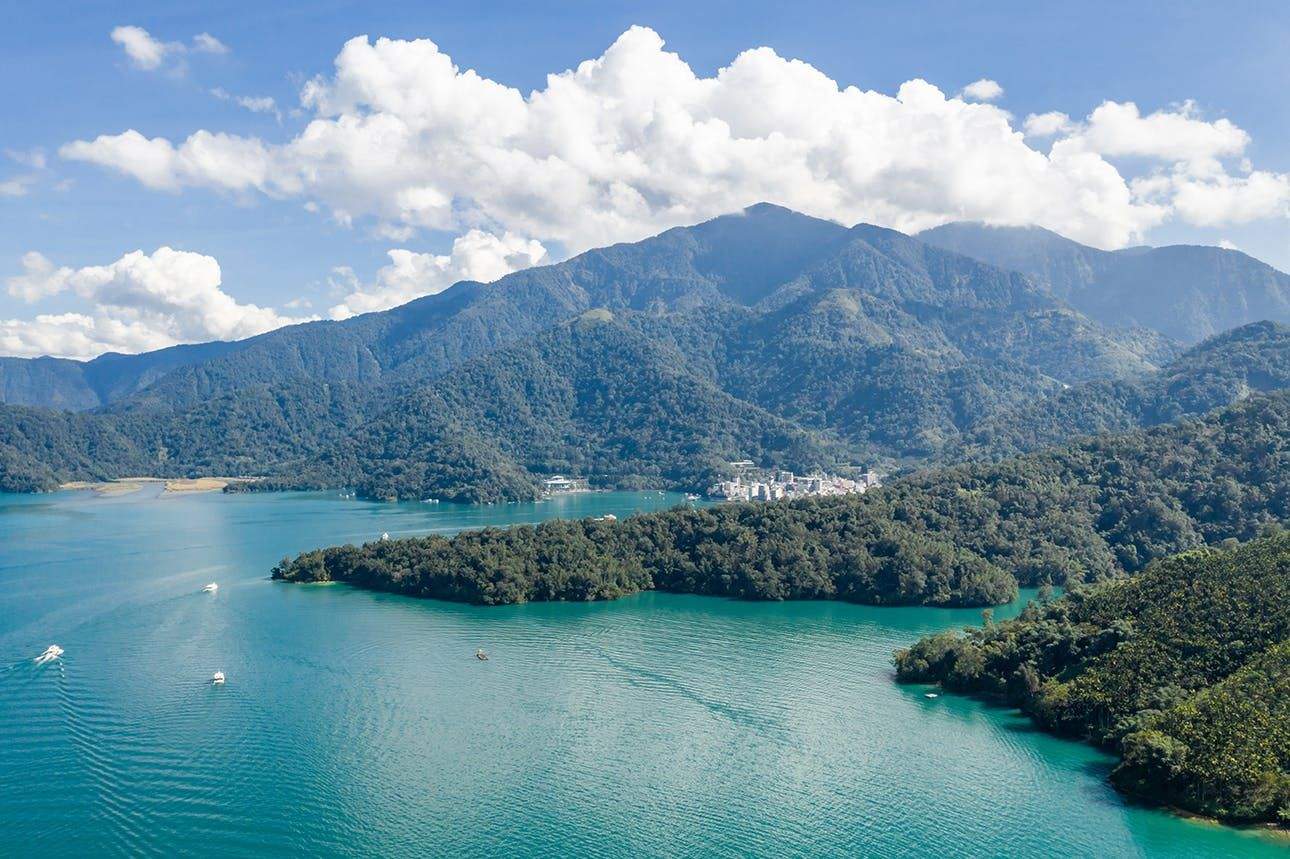 A scenic view of a lake surrounded by forested hills and mountains under a blue sky with scattered clouds.