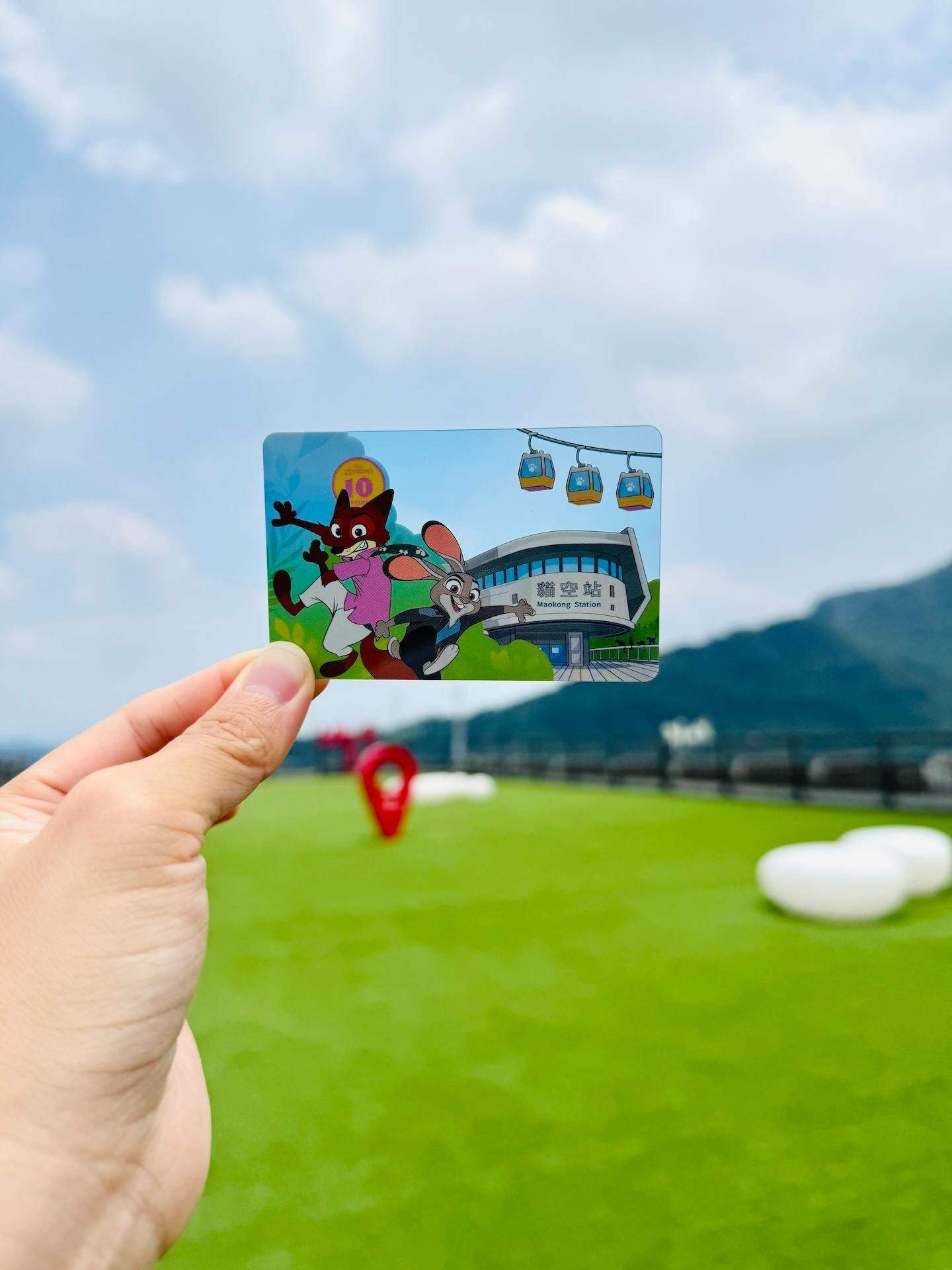 A hand holding a colorful card depicting cartoon characters and a station, with a grassy field and sky in the background.
