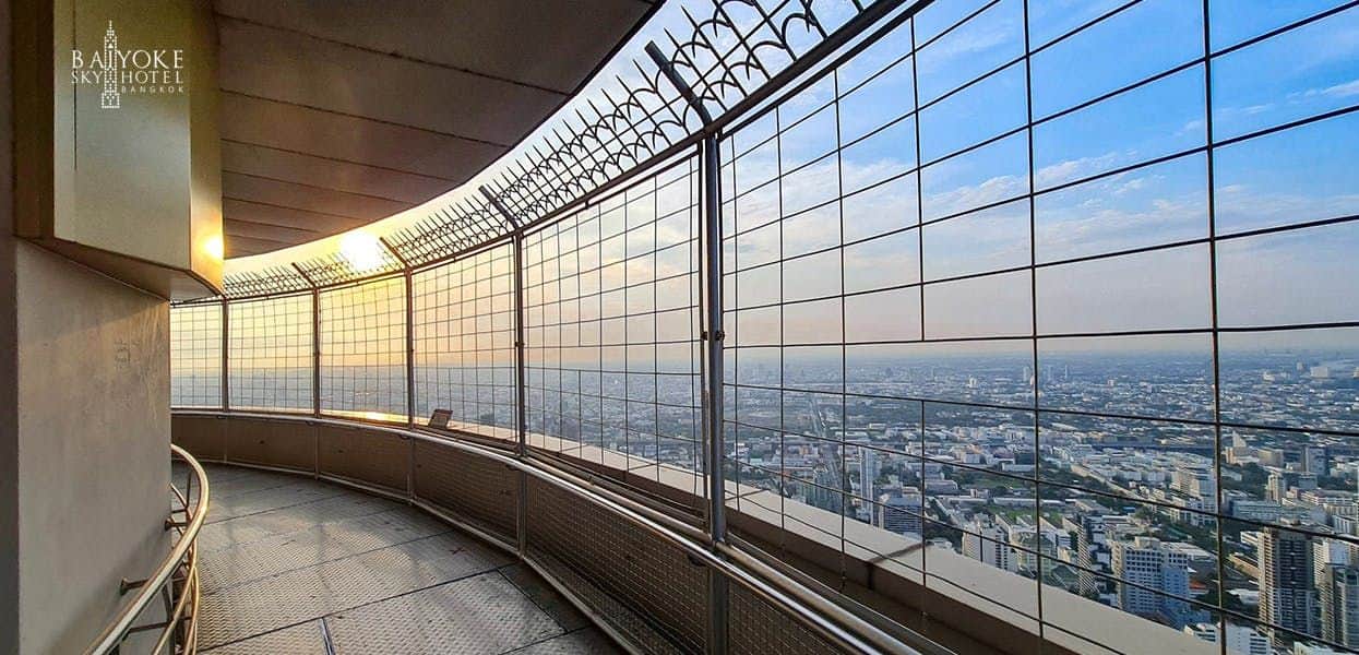 Baiyoke Sky Hotel An outdoor observation deck with metal railings and a wire fence offers a panoramic view of a city skyline at sunset.