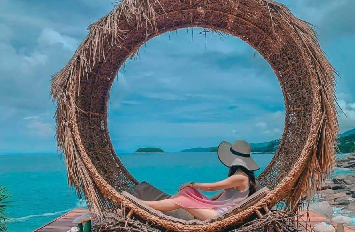 Phuket Rock Beach Scenic Spot A woman in a large sun hat lounges in a circular, woven structure by the sea, with an island and cloudy sky in the background.
