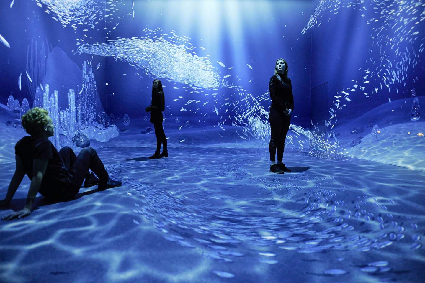 Remastered Three people in a dark, immersive room with projections of underwater scenes, including schools of fish and aquatic structures.