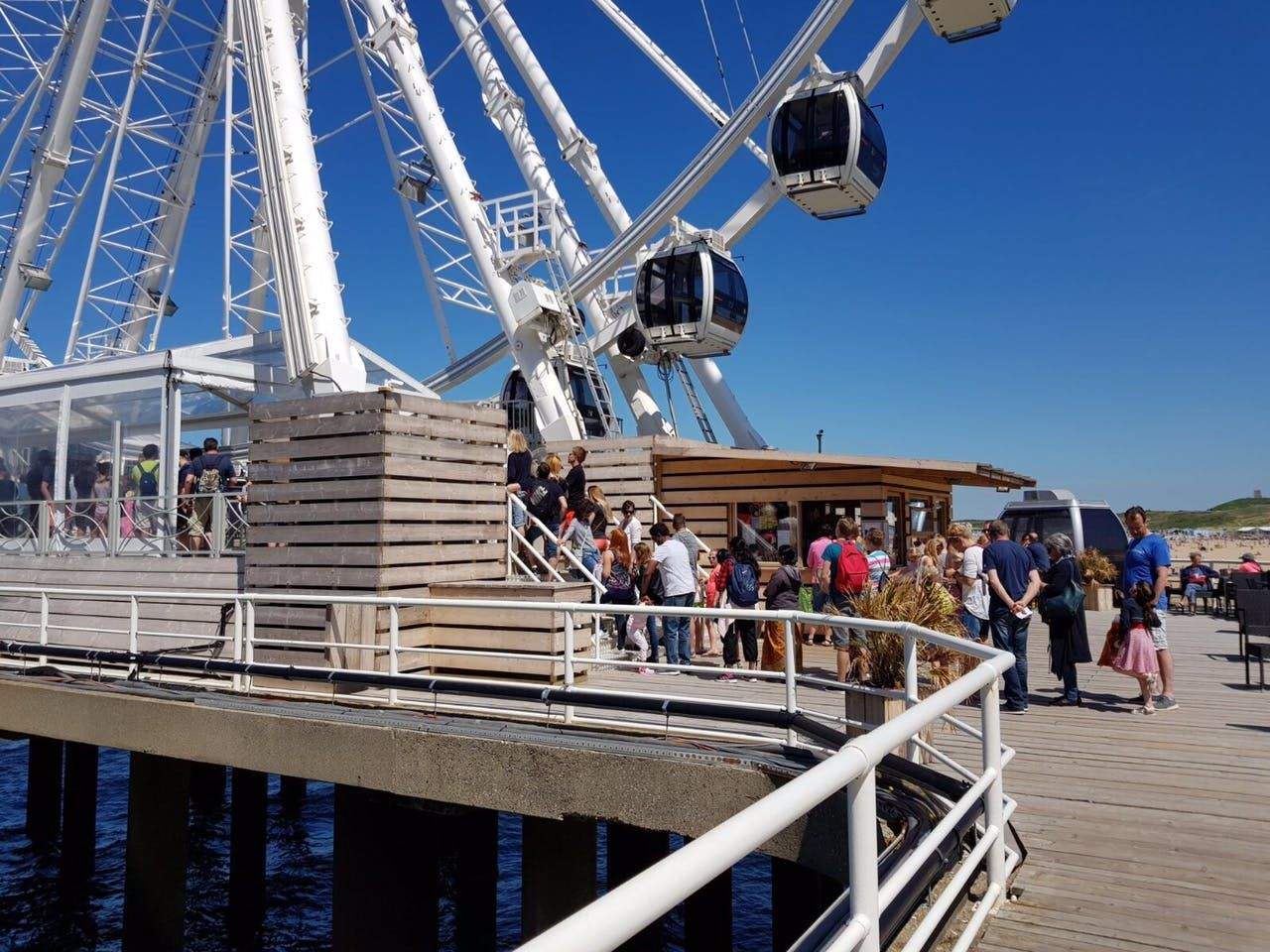 SkyView de Pier People waiting in line at the base of a large Ferris wheel near a wooden building on a sunny day by the water.