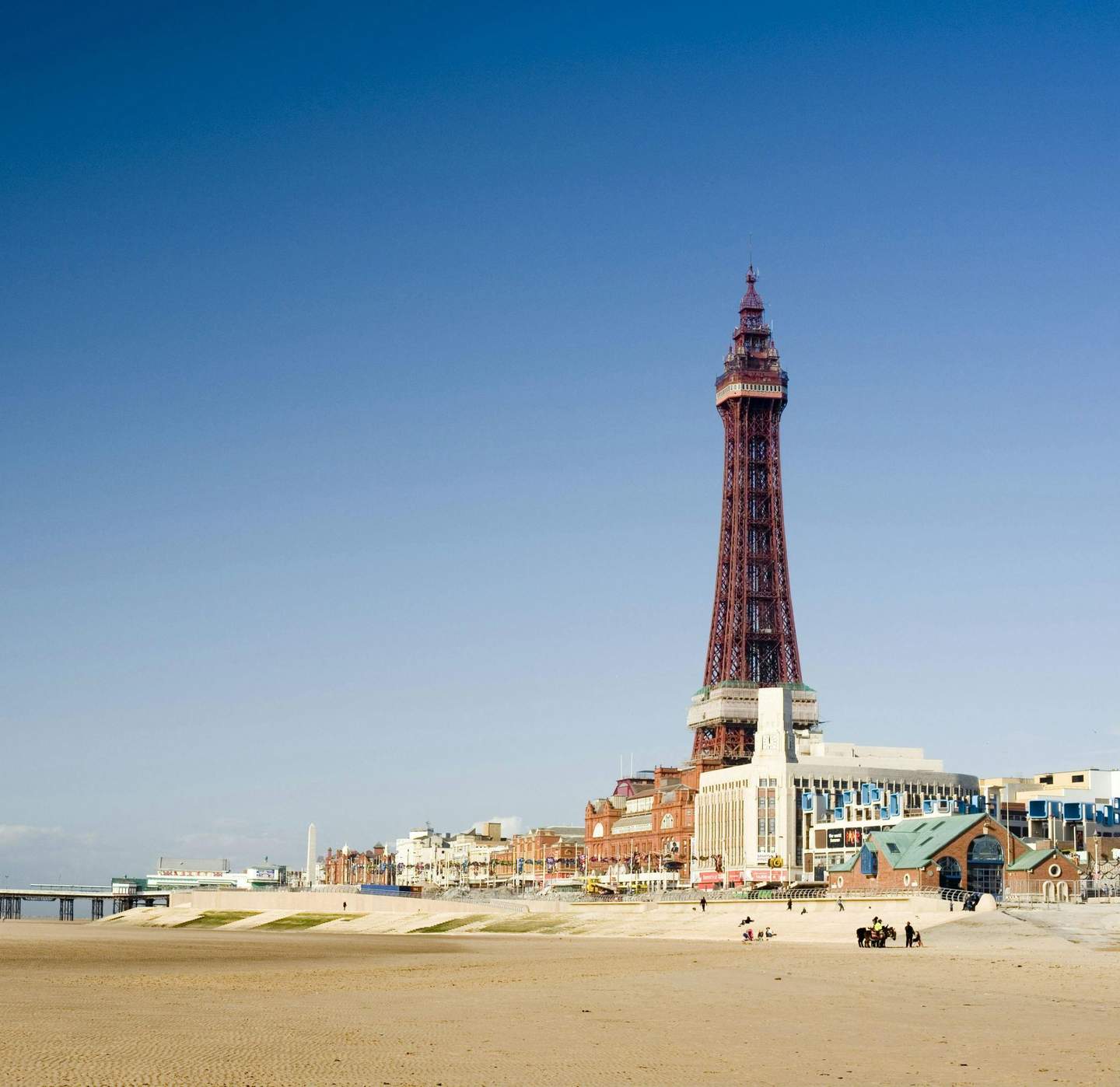 The Blackpool Tower Coastal scene with a tall tower, sandy beach in foreground, and buildings along a promenade under a clear blue sky. Few people visible.