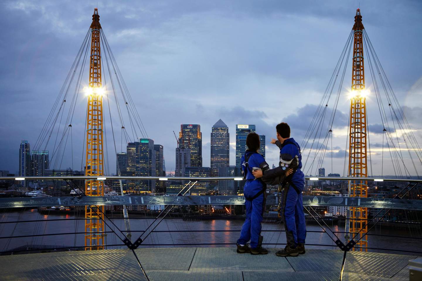 Two people in blue jumpsuits stand on a high platform, overlooking a city skyline at dusk with skyscrapers and a river below.