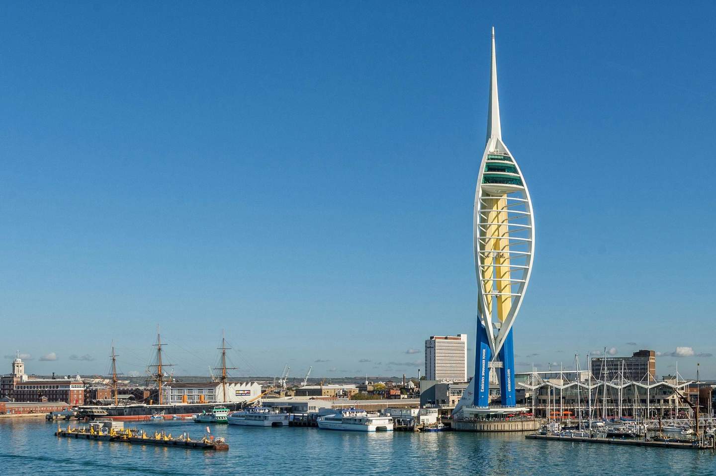 The Spinnaker Tower A tall, modern tower with an observation deck by a waterfront, surrounded by ships, boats, buildings, and a clear blue sky.