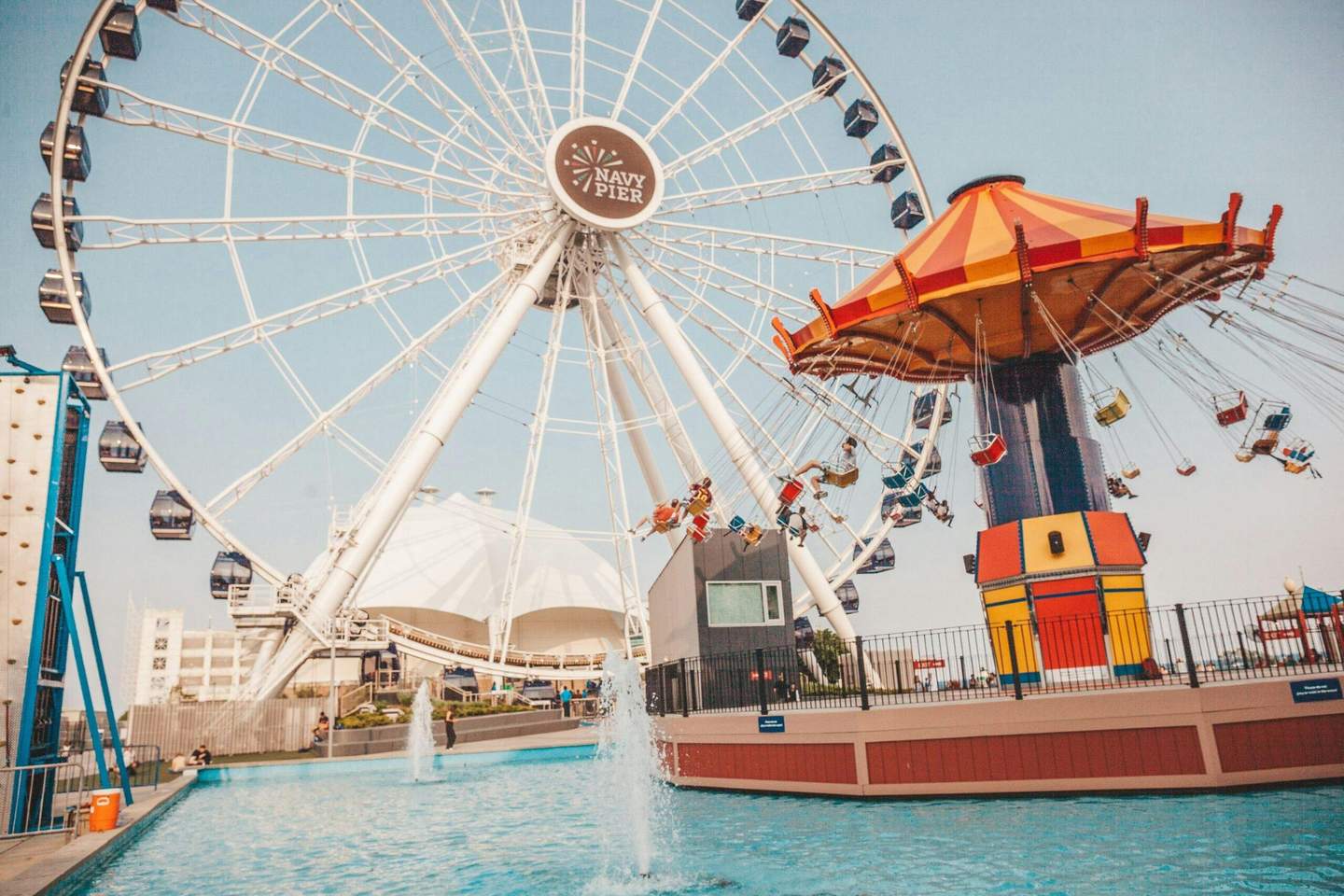 Navy Pier Centennial Wheel A Ferris wheel and a carousel with swings located near a pool with people around, all under a clear sky at Navy Pier.