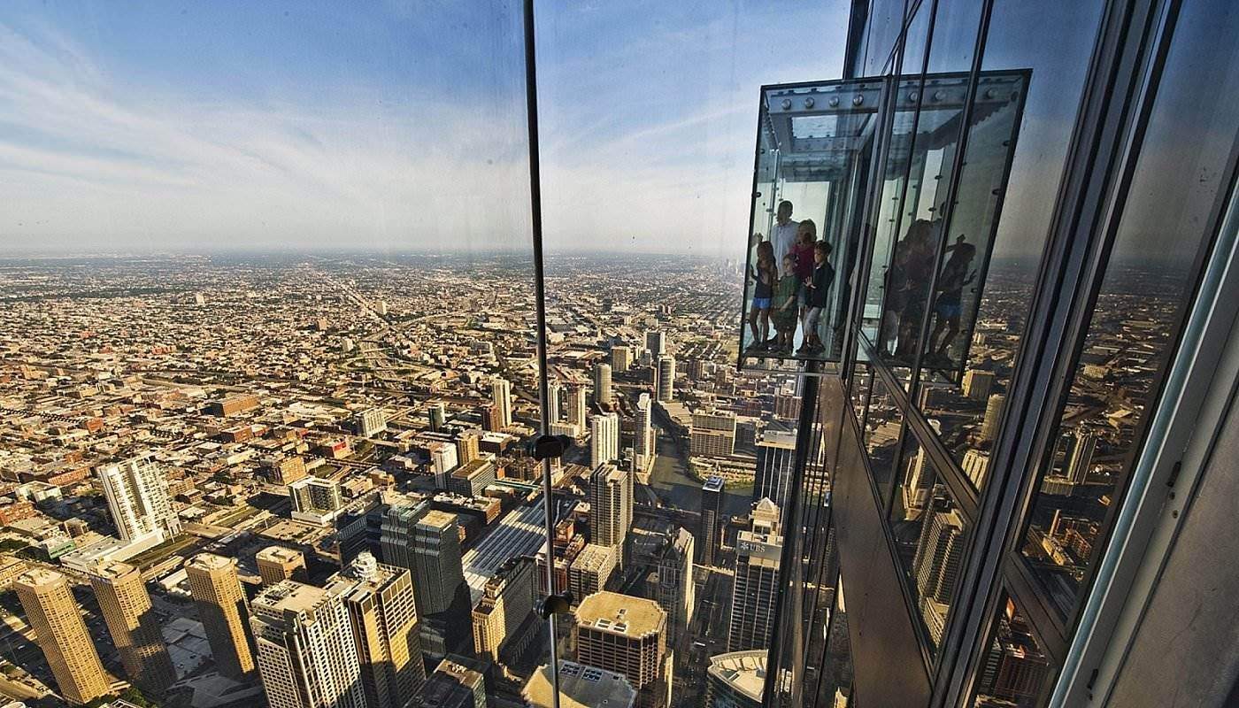People in a glass observation box high above a sprawling cityscape with skyscrapers and distant horizon under a blue sky.