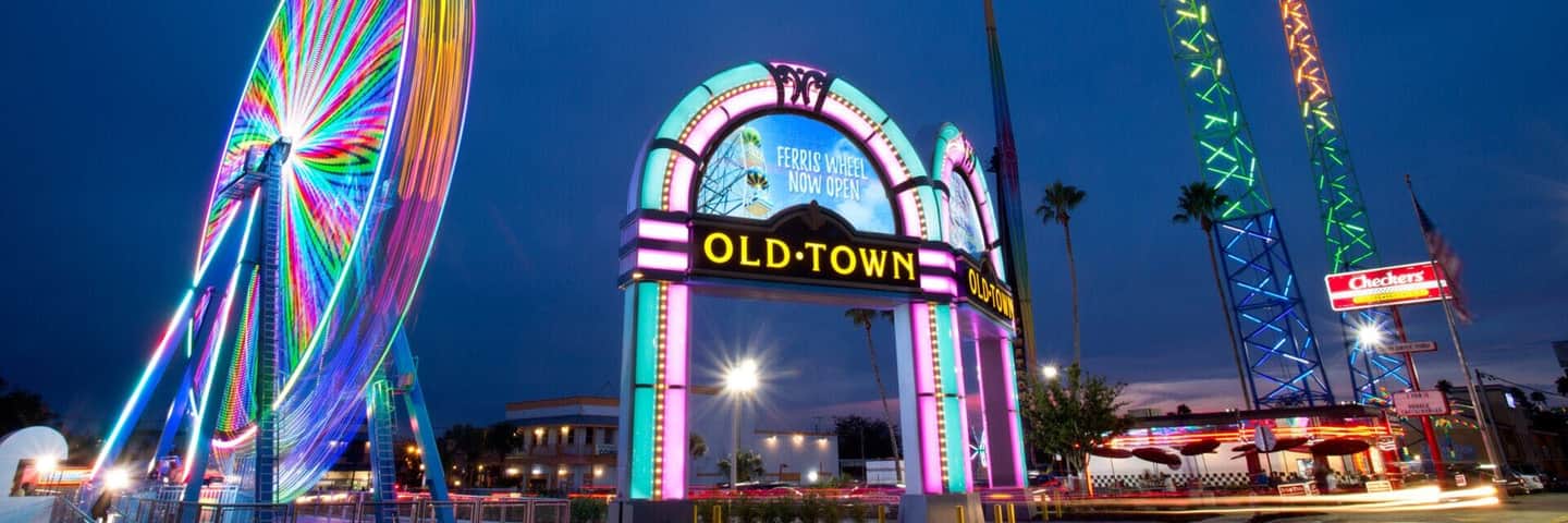 A neon-lit entrance arch reading 'Old Town' at dusk, with a colorful ferris wheel and Checkers sign in the background.