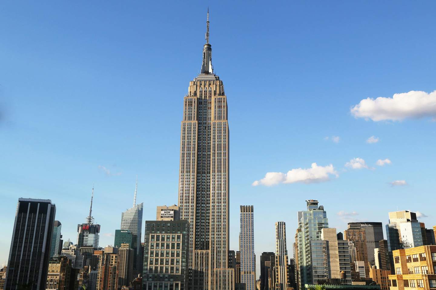 Empire State Building Tall skyscraper with a spire, surrounded by other high-rise buildings under a clear blue sky with a few clouds.