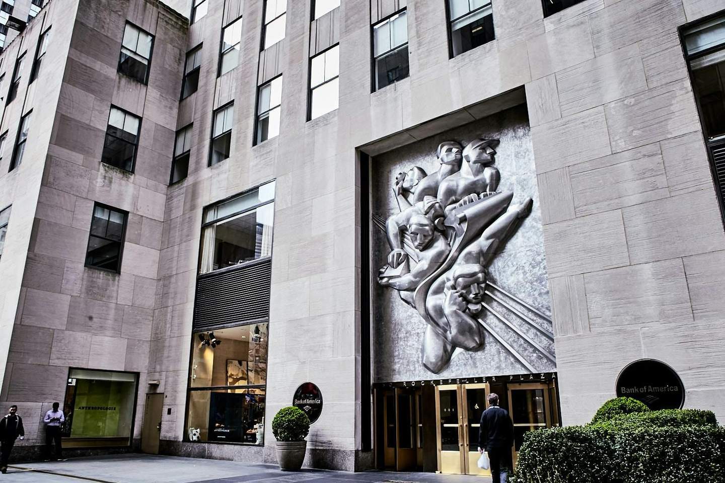 Rockefeller Center Large stone building with high windows, a metal relief sculpture, entrance doors labeled "Rockefeller Plaza," and Bank of America signage.