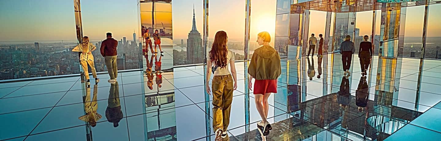 SUMMIT One Vanderbilt People stand and walk on a mirrored observation deck with a view of a city skyline at sunset.