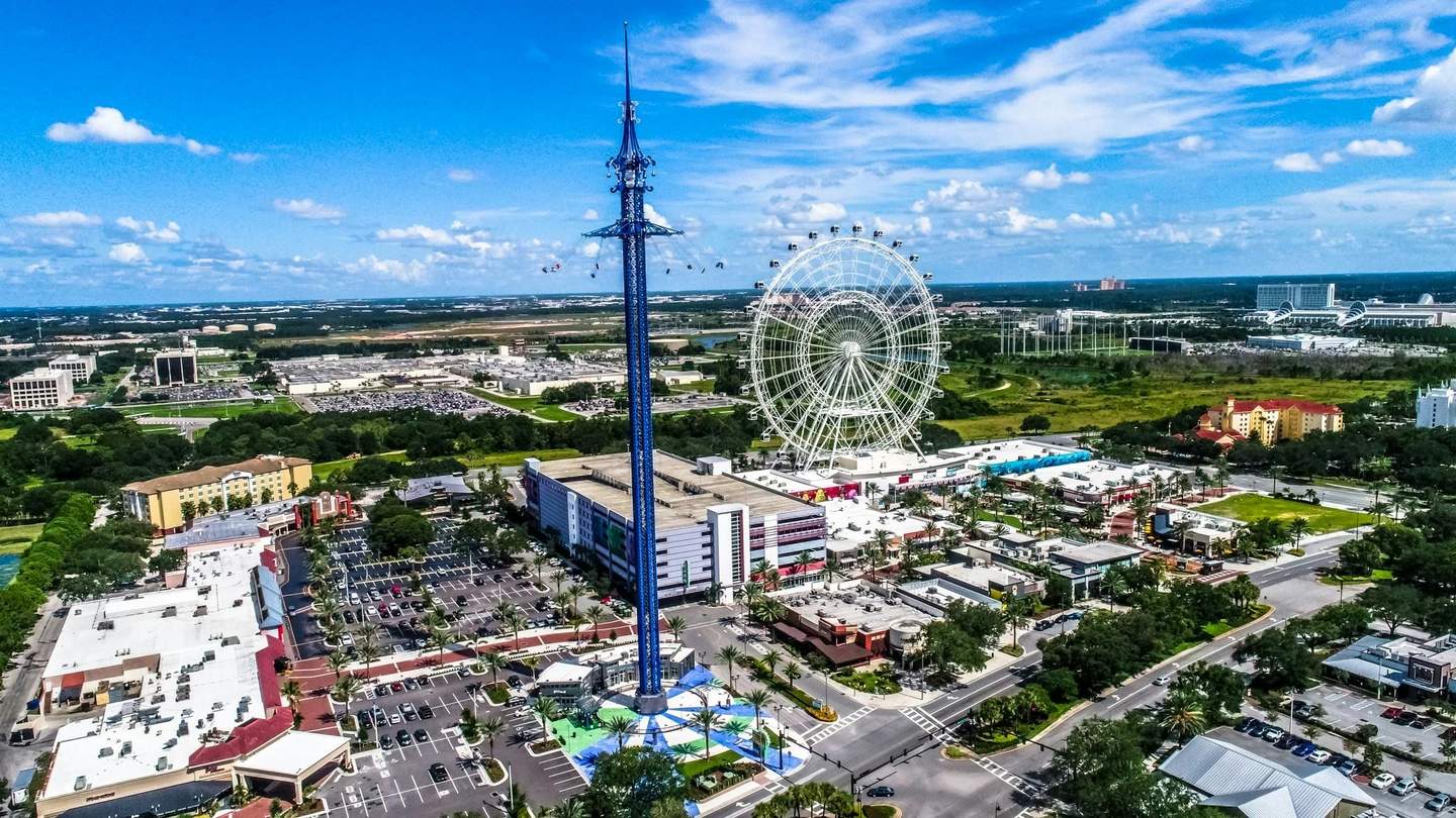 The Orlando Eye Aerial view of an amusement park with a tall swing ride, Ferris wheel, parking lots, buildings, and surrounding greenery under a blue sky.