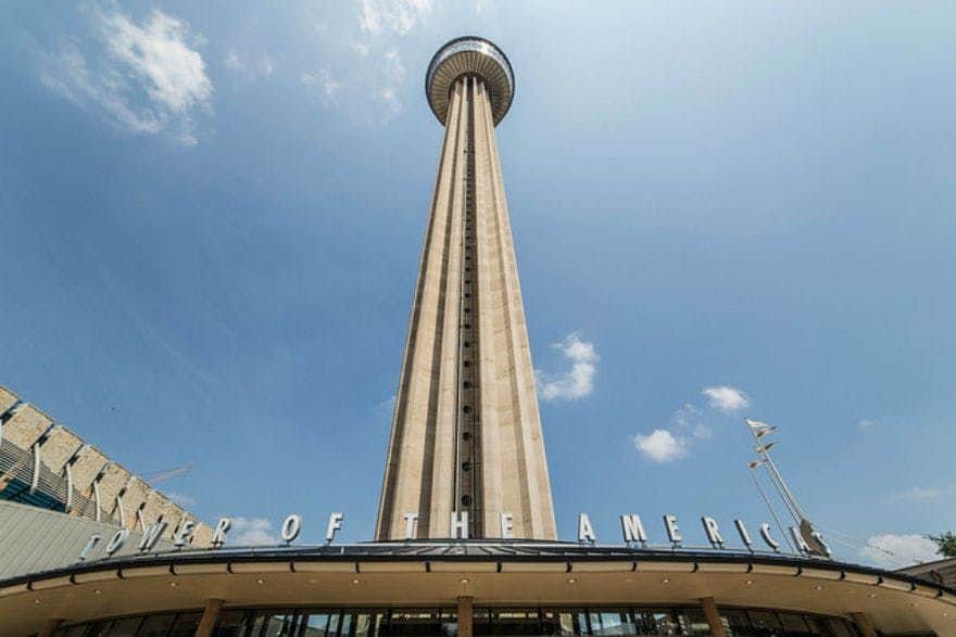 Tall observation tower with a circular deck, labeled "Tower of the Americas," under a clear blue sky with a few clouds.