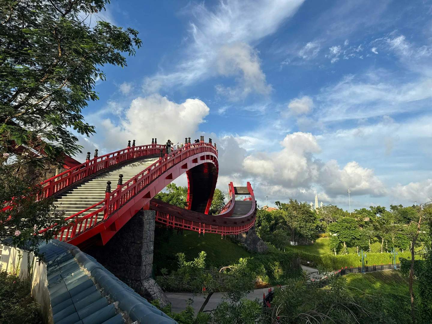 Sun World Ha Long Czerwony łukowaty most dla pieszych z balustradami rozpościera się na zielony park pod błękitnym niebem z chmurami.