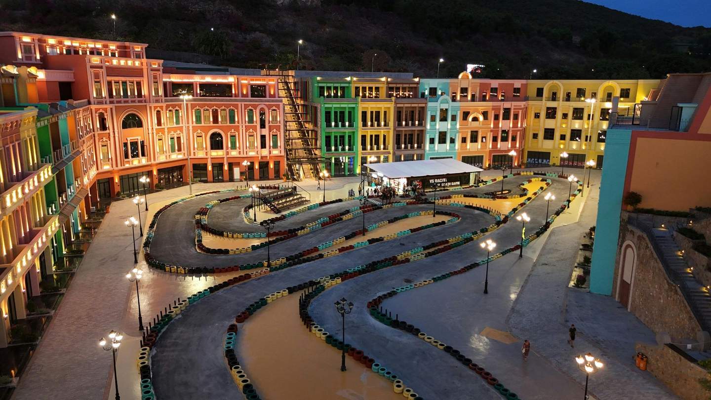 Uma pista de kart iluminada, rodeada por edifícios coloridos e animados ao anoitecer, com algumas pessoas a passear nas proximidades.