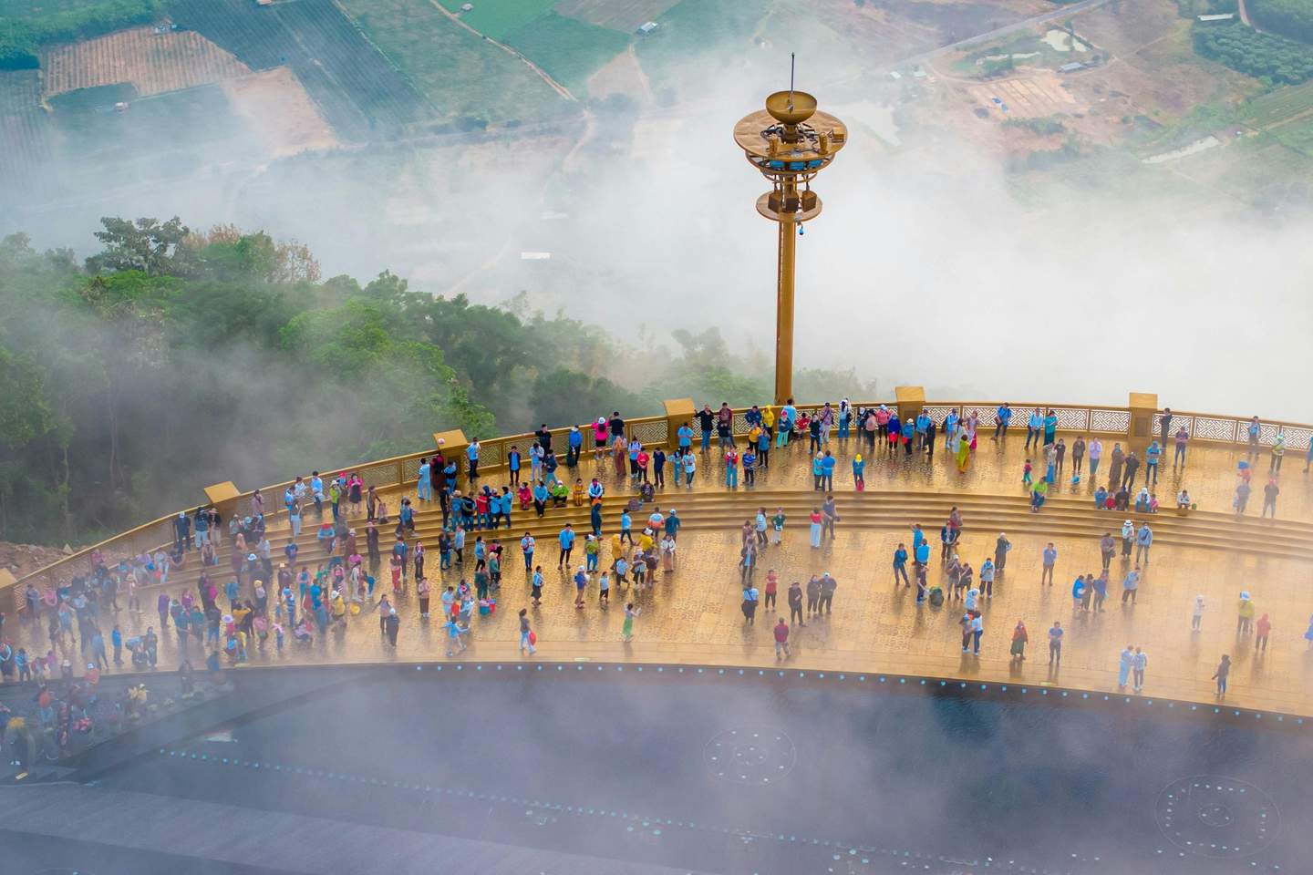 People standing and walking on a large golden platform with ornamental railings, surrounded by fog and overlooking a green landscape.