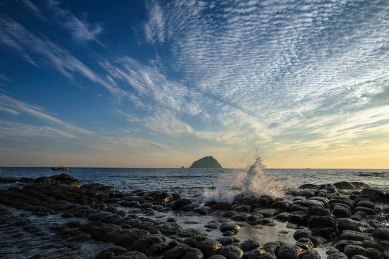 台北趣味探索通票: 2日景点通票 海浪沿着海岸线冲击着圆形的岩石,远处有一个岛屿,天空中充满了零星的云层。