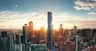 A city skyline at sunrise featuring a tall, modern skyscraper with a distinctive design surrounded by other buildings.