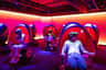 People seated in pod-like chairs, wearing virtual reality headsets, in a room with vibrant pink and purple lighting.