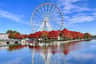 A large Ferris wheel is reflected in a calm lake, surrounded by trees with vibrant red foliage under a blue sky.
