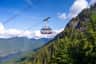 A red cable car labeled "Grouse Mountain" is suspended above a forested mountain landscape under a blue sky.