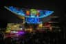 A nighttime scene showing a building with colorful light projections of cartoon bears and fireworks, surrounded by a crowd.