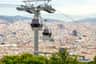 Two cable cars travel over a densely built city with mountains in the background and greenery in the foreground.