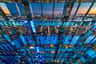Mensen die een stadslandschap bekijken vanuit een glazen structuur met meerdere reflecties en neonblauwe verlichting.